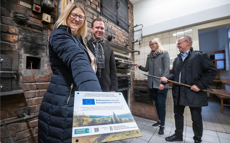 Barbara Smith (Geschäftsführerin von „Leader“ Heckengäu) überreicht die Plakette über die Förderung an den Deckenpfronner Bürgermeister Dennis Mews, während im Hintergrund Kämmerin Kathrin Luz und Ralph Süßer (erster stellvertretender Bürgermeister und Gemeinderat) die Elektroöfen begutachten. Diese ersetzen den alten Steinofen auf der rechten Seite.GB-Foto: Vecsey